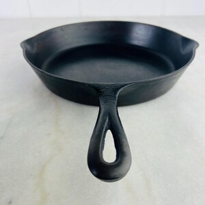 May include: A black cast iron skillet with a long handle featuring a hole for hanging. The skillet has a wide, shallow cooking surface and two small spouts. This cookware is ideal for frying and searing.