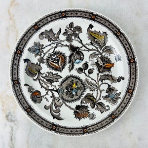 May include: A white ceramic plate with a black and brown floral design. The plate has a decorative border with a repeating pattern.