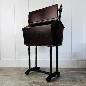 May include: A dark brown wooden sewing box with a hinged lid and a deep storage compartment. The box is supported by four slender, turned legs connected by a horizontal stretcher bar at the base, featuring a sturdy, elevated rectangular design.