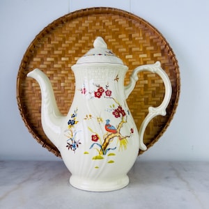 Könnte beinhalten: Cremefarbene Keramik-Kaffeekanne mit Deckel, mit einem Blumen- und Vogeldesign in Rot, Blau und Gelb. Die Kanne hat einen gebogenen Ausguss und Griff und steht vor einem gewebten Korbhintergrund.