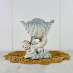 May include: A vintage porcelain figurine of a cherub holding a seashell-shaped bowl. The cherub has blonde hair, blue wings, and red lips. The seashell is white with blue accents. The figurine is on a brown doily.