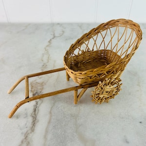 May include: A small, brown wicker wheelbarrow with a single wheel. The wheelbarrow is made of woven wicker and has a rounded, oval-shaped basket.