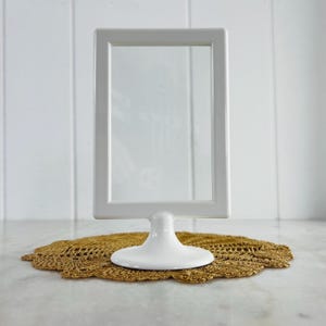 May include: A white, rectangular picture frame with a clear glass insert, mounted on a pedestal base. The frame is displayed on a gold-colored, crocheted doily. The frame is approximately 6 inches tall.