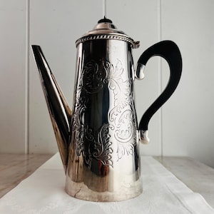 Puede incluir: Una cafetera de metal plateado con un cuerpo cilíndrico alto y patrones de volutas grabados. Incluye un pico largo y cónico, una tapa con bisagras con un pequeño remate y un asa negra curva sujeta con soportes de metal plateado.