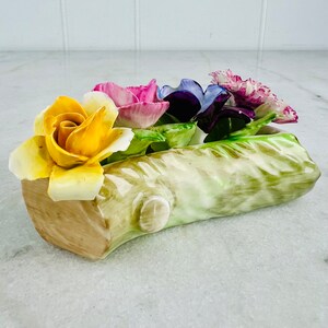 Puede incluir: Un arreglo floral de porcelana con una rosa amarilla, una rosa rosa, una violeta morada y un clavel rosa. Las flores están dispuestas en una base con forma de tronco.