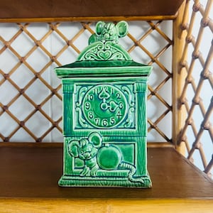 Puede incluir: Un recipiente de cerámica verde con forma de reloj con un diseño de ratón. La esfera del reloj presenta números romanos. Una figura de ratón está en la parte superior y otra en la parte delantera. El recipiente está sobre una estantería de madera.