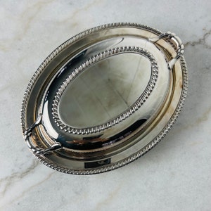 Könnte beinhalten: Silberne ovale Servierplatte mit Deckel. Die Platte hat einen perlenbesetzten Rand und zwei Griffe. Der Deckel hat einen transparenten Glaseinsatz.