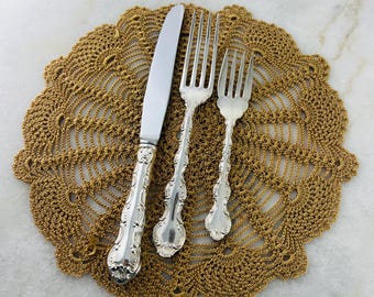 May include: A set of three silver-plated utensils, including a knife and two forks, arranged on a gold crochet doily.
