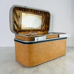 May include: A vintage tan vanity case with a silver trim and handle. The interior features a mirror and patterned lining. The case is rectangular with rounded corners and a black stripe detail. The case is open, revealing the interior.