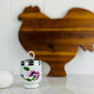 May include: A white ceramic egg cup with a floral design and a silver lid. The egg cup is sitting on a white countertop next to a white egg and a wooden chicken-shaped chopping board.