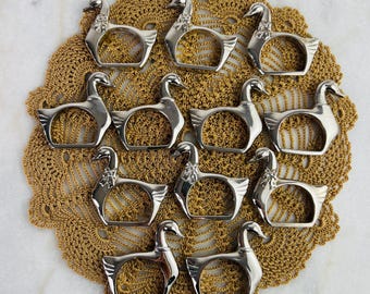 Può includere: Una collezione di dodici anelli portatovaglioli in metallo argentato a forma di anatra. Gli anelli sono disposti su un centrino dorato all'uncinetto. Le anatre hanno dettagli, tra cui becchi, occhi e ali. Lo sfondo è una superficie di marmo bianco.