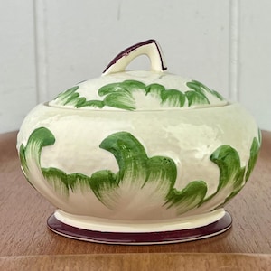 Puede incluir: Un azucarero de cerámica blanca con tapa, decorado con diseños florales verdes y morados. El tazón tiene una forma redondeada y un borde festoneado.