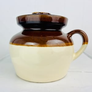 Könnte beinhalten: Eine braune und weiße Keramik-Dose mit Deckel und Henkel. Die Dose hat eine braune Glasur auf der oberen Hälfte und eine weiße Glasur auf der unteren Hälfte.