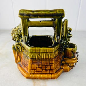 Pode incluir: Um vaso de cerâmica em forma de poço dos desejos, com um esquema de cores castanho e verde. O poço apresenta uma base em forma de tijolo, uma estrutura de madeira e um pequeno balde pendurado por uma corrente. A palavra "WISHING" está escrita na frente.