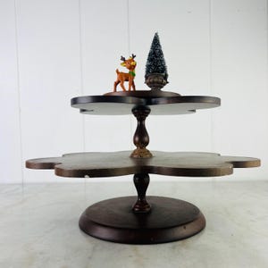 Puede incluir: Una bandeja de madera marrón oscuro de dos niveles con una base redonda y un borde decorativo festoneado. Una pequeña figura de reno y un árbol de Navidad en miniatura se exhiben en el nivel superior. La bandeja es adecuada para decoraciones.