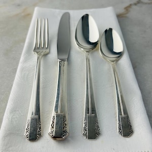 May include: A set of four silver-coloured cutlery pieces: a fork, a knife, and two spoons. The utensils are arranged on a white cloth. Each piece has a decorative design on the handle.