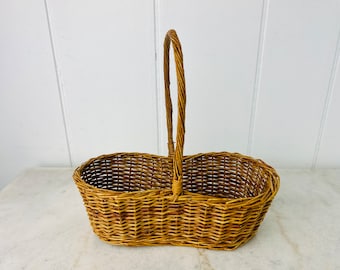 Könnte beinhalten: Ein ovaler Weidenkorb mit einem zentralen Griff. Der Korb hat einen geteilten Innenraum. Der Griff ist hoch und gewölbt, aus dem gleichen geflochtenen Material wie der Korb. Der Korb hat eine natürliche braune Farbe.
