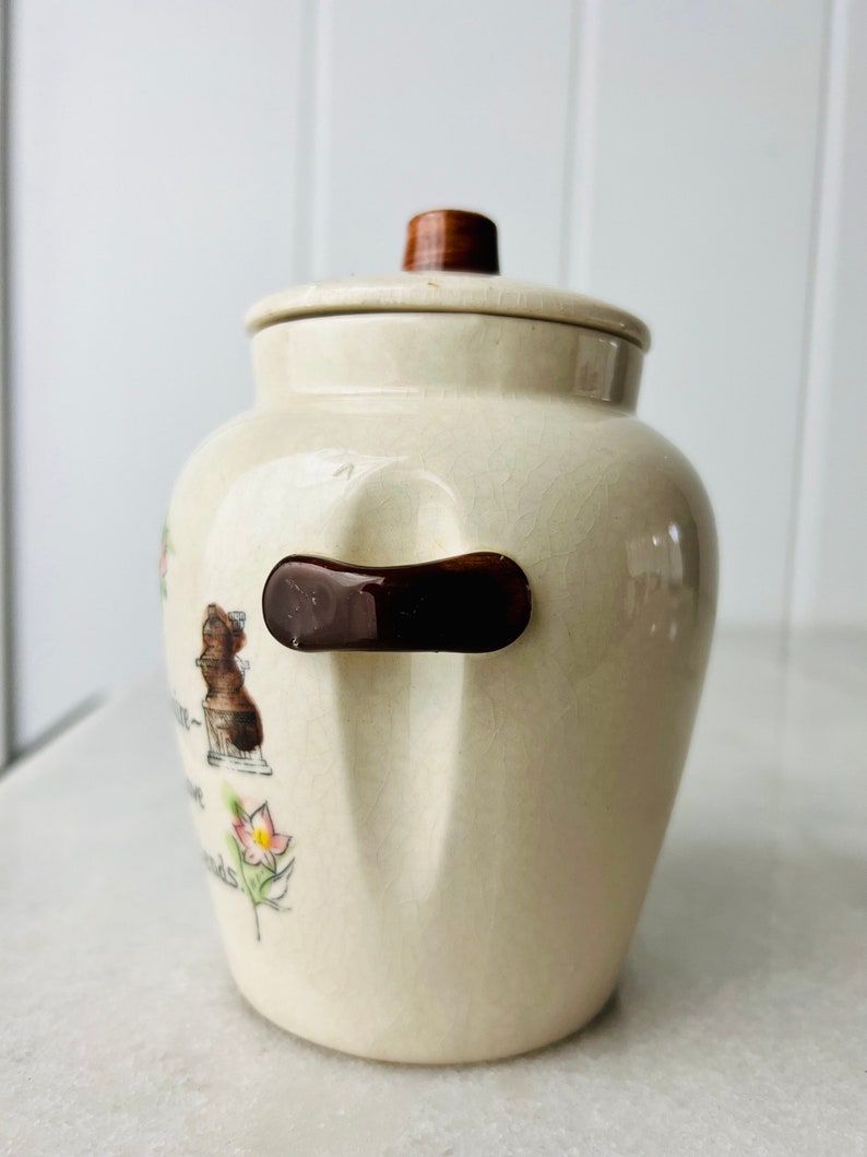 K&ouml;nnte beinhalten: Eine wei&szlig;e Keramik-Keksdose mit braunem Deckel und Henkel. Die Dose hat ein Blumenmuster und den Text "A Little Bit of Love Goes a Long Way".