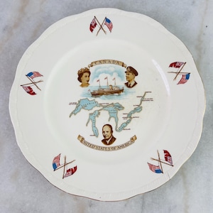 May include: Cream-coloured decorative plate with a scalloped edge and gold trim. The plate features a map of the Great Lakes, portraits, and the flags of the United States and Canada. The plate has the words "Canada" and "United States of America".