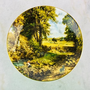 May include: A round porcelain plate with a gold rim features a painting of a rural landscape. The scene depicts a winding path through a field with trees, a stream, and a group of sheep. The painting is in a traditional style with muted colors.