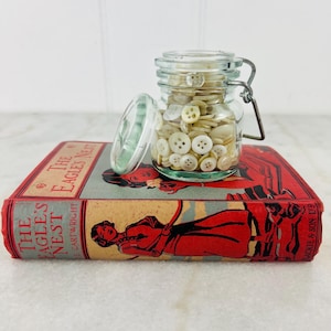 May include: A glass jar filled with white and cream colored buttons sits on top of a red and blue book titled "The Eagle's Nest" by E. Cartwright.