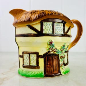 Puede incluir: Una jarra de cerámica con forma de cabaña con techo de paja, puerta de madera marrón y ventanas con cristales en forma de diamante. La cabaña está pintada en tonos amarillos, marrones y verdes.