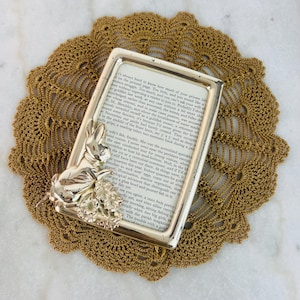 May include: A silver-colored picture frame with a detailed bunny and floral design on the corner, resting on a gold-colored crocheted doily. The frame holds a page of printed text. The doily has a circular shape.