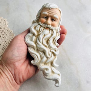 May include: A ceramic Santa Claus head with a long, flowing white beard and rosy cheeks. The Santa figure has a detailed face with a slight smile and is likely a decorative item for the holiday season.