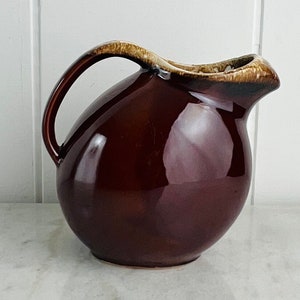 Peut inclure: Un pichet en céramique brun avec un corps large et arrondi et une anse. Le pichet est recouvert d'un glaçage brun avec un bord brun clair.