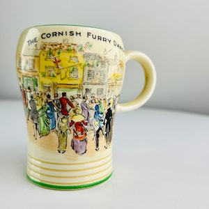 Puede incluir: Una taza de cerámica de color crema con un borde verde. La taza presenta una ilustración colorida de una escena callejera con gente bailando. El texto "The Cornish Furry Dance" está impreso en la taza.