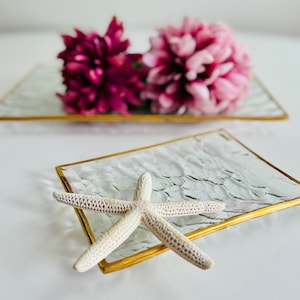 May include: A white starfish on a clear glass tray with a gold rim. The tray is sitting on a white surface.