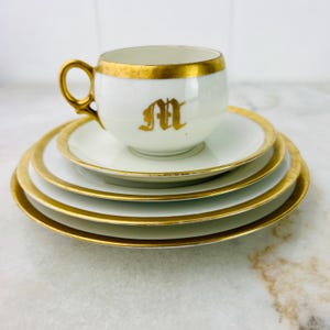 Puede incluir: Un juego de taza y platillo de té blanco con ribete dorado. La taza de té tiene una inicial 'M' dorada en la parte delantera. El juego está apilado sobre dos platillos adicionales.