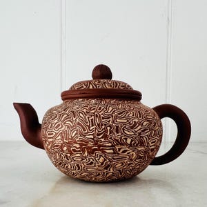 Op de afbeelding: Een ronde keramische theepot met een donkerbruin en crèmekleurig gemarmerd patroon op het lichaam en het deksel. De theepot heeft een effen donkerbruine tuit, een bijpassend gebogen handvat en een kleine, ronde knop op het deksel.