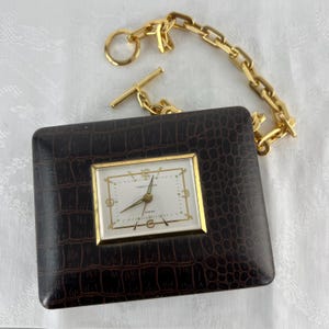 May include: A vintage travel clock with a brown, faux-crocodile skin case and a gold-tone frame. The clock face is white with black numerals and hands. A gold chain is attached to the case.