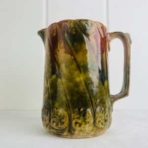 May include: A vintage ceramic jug with a textured, swirling design in shades of green, brown, and gold. The jug has a handle and a spout, and the base features embossed floral patterns. The overall aesthetic is rustic and antique.