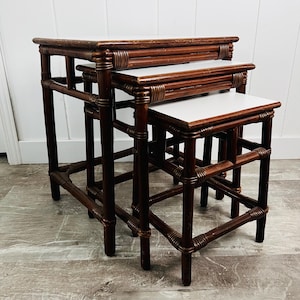 May include: A set of three nesting tables featuring dark brown bamboo-style frames with wrapped detailing at the joints and smooth, light-colored rectangular tops. The tables are arranged in a cascading formation, showing varying heights and sizes.