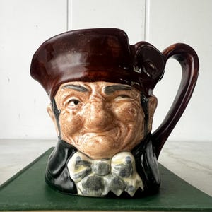 Peut inclure: Une cruche en céramique en forme de personnage portant un tricorne marron, un manteau noir et une cravate blanche à motifs. Le visage présente des textures de peau détaillées, un nez proéminent et une expression subtile avec un œil plissé.