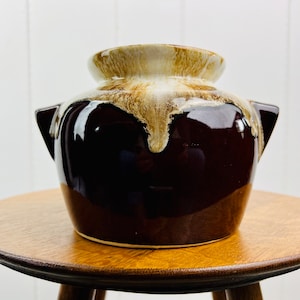 Könnte beinhalten: Ein brauner und weißer glasierter Keramiktopf mit zwei Griffen. Der Topf hat ein Tropf-Glazurmuster und steht auf einem Holztisch.