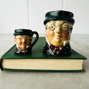 May include: Two ceramic character mugs featuring a person wearing a dark green hat, round glasses, and a dark coat with a red bowtie. The mugs are glazed in shades of tan, green, and red, depicting a detailed facial expression with rosy cheeks.