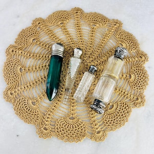 Puede incluir: Cuatro frascos de perfume antiguos con tapones plateados descansan sobre un tapete dorado de ganchillo. Los frascos varían en forma y tamaño, incluyendo un frasco verde oscuro y varios frascos de vidrio transparente.