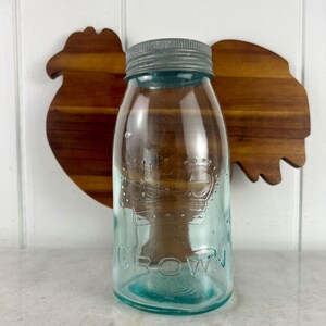 Op de afbeelding: Een vintage aqua blauwe glazen pot met een zilveren deksel. De pot heeft het woord "CROWN" gegraveerd op de bodem en een kroonontwerp gegraveerd aan de zijkant.