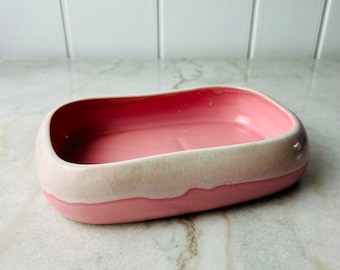 May include: A pink ceramic soap dish with a white rim. The dish has a rectangular shape with rounded edges.