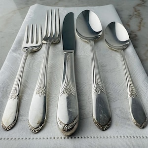 May include: A collection of five silver-coloured cutlery items, including forks, a knife, and spoons, arranged on a white cloth. The silverware features ornate detailing on the handles and a decorative border. The spoons and forks are curved, and the knife has a straight blade.
