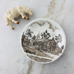 May include: A white ceramic saucer with a brown and white illustration of a village scene. The saucer has the text "Toll House Upper Canada Village" printed on the bottom.