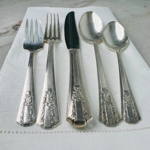 May include: Silverware set including two forks, a knife, and two spoons. The utensils have a decorative floral design on the handles and are arranged on a white cloth. The knife has a black blade.