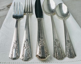 Könnte beinhalten: Silbernes Besteck-Set mit zwei Gabeln, einem Messer und zwei Löffeln. Das Besteck hat ein dekoratives Blumendesign an den Griffen und ist auf einem weißen Tuch angeordnet. Das Messer hat eine schwarze Klinge.