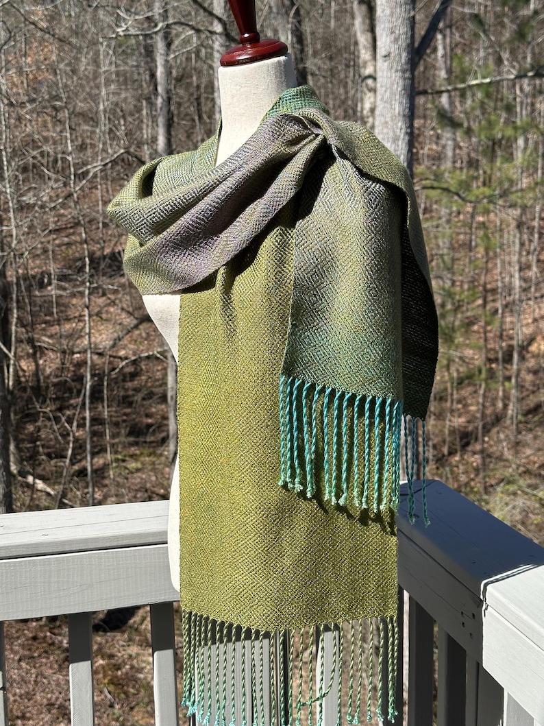 May include: A green and blue woven scarf with a fringe. The scarf is draped over a mannequin.