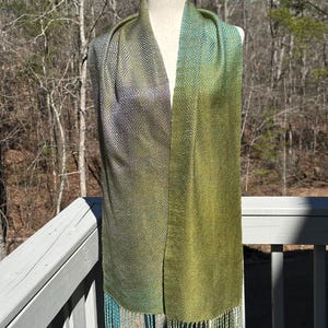 May include: A green and blue silk scarf with a geometric pattern and fringe. The scarf is displayed on a mannequin against a backdrop of trees.