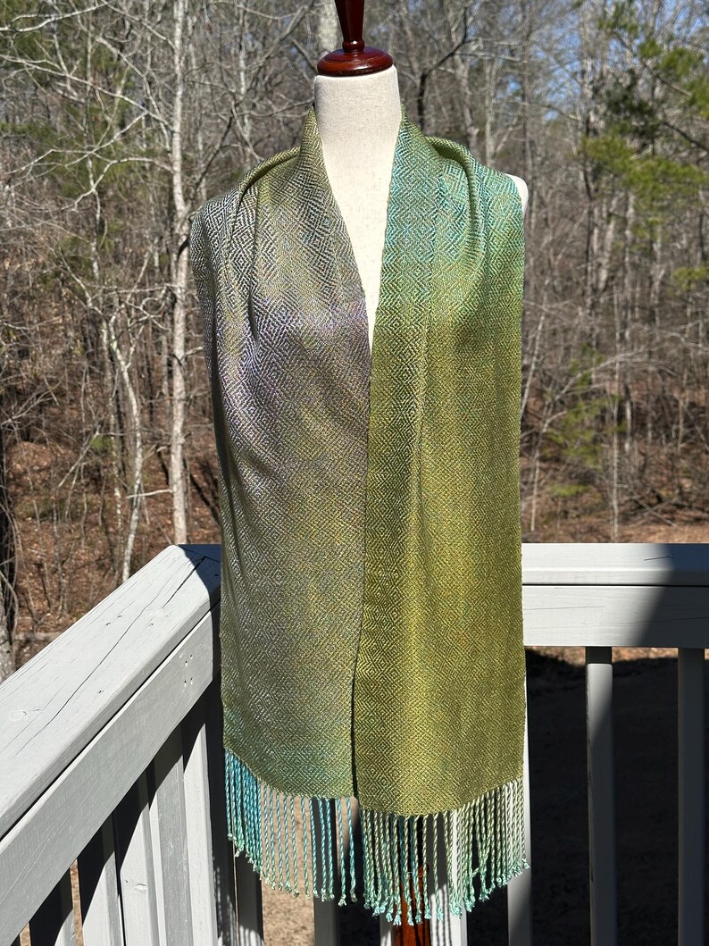 May include: A green and blue silk scarf with a geometric pattern and fringe. The scarf is displayed on a mannequin against a backdrop of trees.