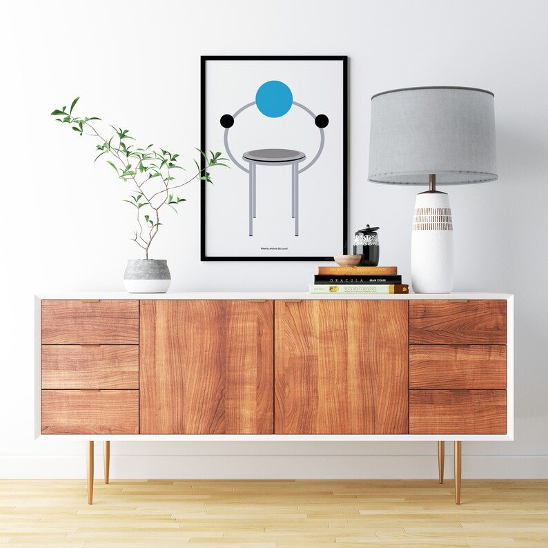 K&ouml;nnte beinhalten: Ein wei&szlig;er Sideboard im Mid-Century-Modern-Stil mit drei Schubladen und zwei T&uuml;ren. Das Sideboard hat eine Holzmaserung und goldene Beine. Ein gerahmtes Bild eines Stuhls mit einem blauen Kreis dar&uuml;ber befindet sich an der Wand &uuml;ber dem Sideboard. Eine Lampe mit einem grauen Schirm und einem wei&szlig;en Fu&szlig; steht auf dem Sideboard. Auf dem Sideboard liegen B&uuml;cher und ein kleines schwarzes Objekt.